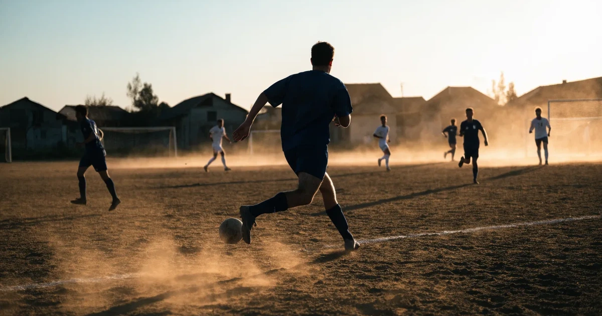 Ein sandiger Fußballplatz am Rand einer Stadt in warmem Abendlicht, Staub in der Luft und zwei Tore in der Ferne.