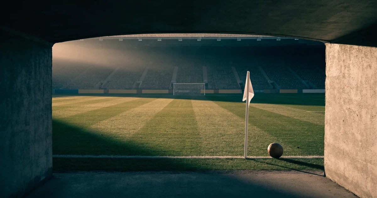 Blick durch ein Stadiontor auf einen grünen Rasen im Abendlicht, ein Ball ruht an der Eckfahne, die Tribünen sind leer.