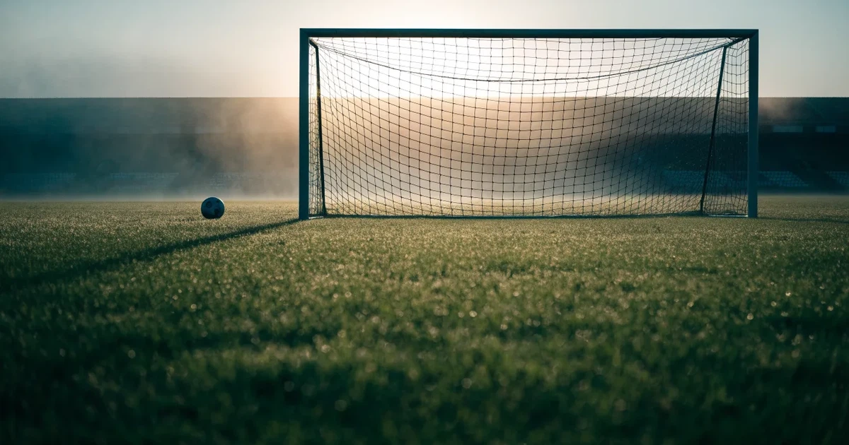 Ein Fußballtor mit gespanntem Netz steht auf einem leeren Rasenplatz, davor ein einzelner Ball, Morgendunst liegt über dem Feld.