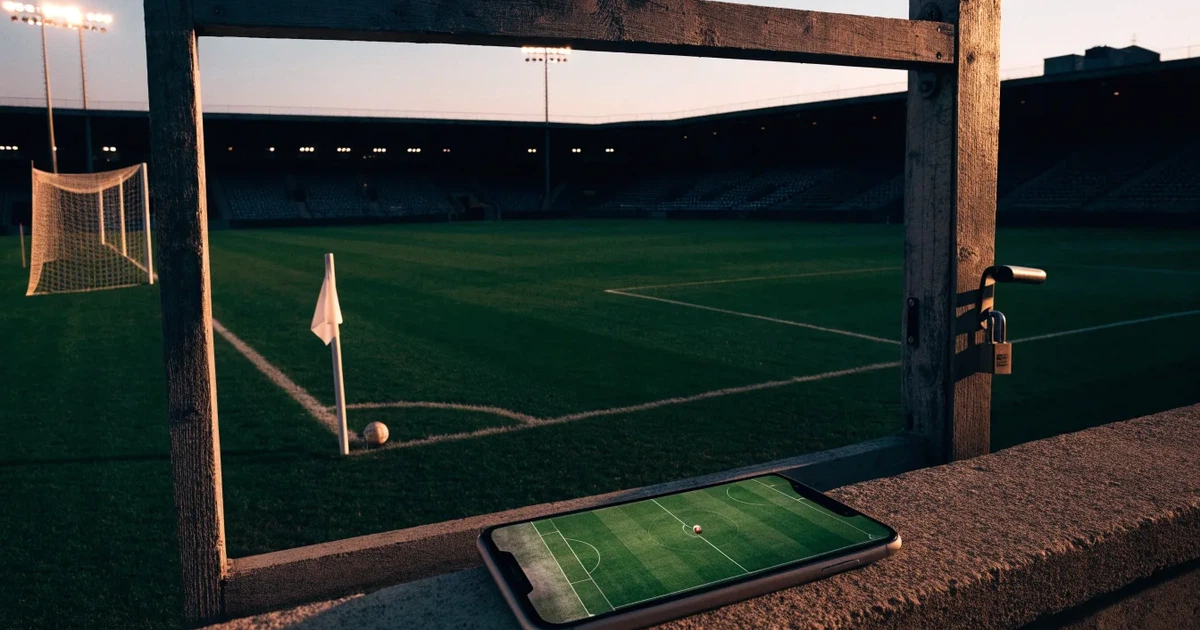 Blick durch ein Stadiontor auf einen grünen Rasen im Abendlicht, ein Ball liegt an der Eckfahne, die Ränge sind leer.