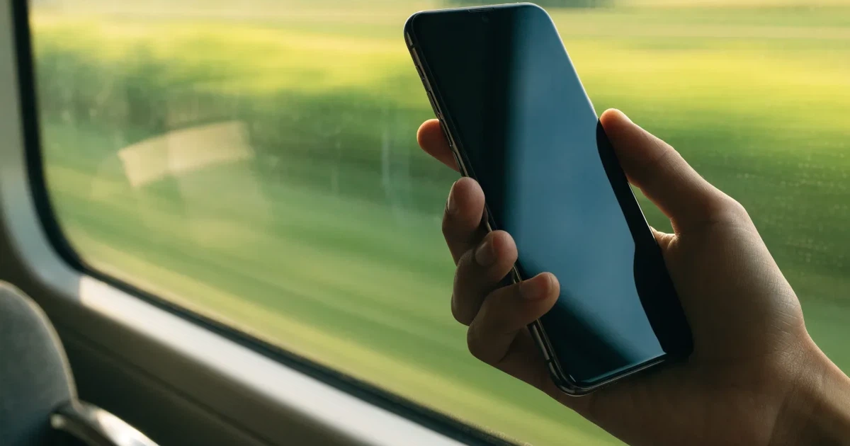 Eine Hand hält ein Smartphone mit dunklem Display in einem Zug, am Fenster ziehen verschwommen Felder vorbei.