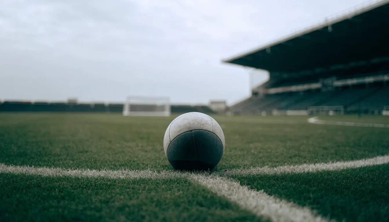 Ein Fußball liegt auf dem Anstoßpunkt eines leeren Rasenplatzes, im Hintergrund eine ruhige Tribüne unter bedecktem Himmel.
