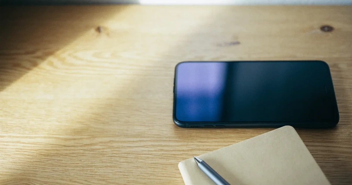 Ein Smartphone liegt neben einem Notizbuch und einem Kugelschreiber auf einem hellen Schreibtisch, das Display ist ausgeschaltet.