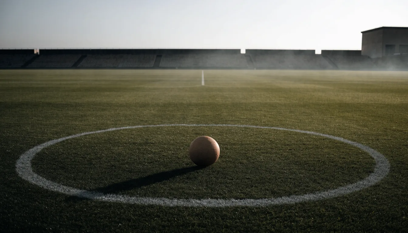 Ein Fußball liegt auf dem Anstoßpunkt eines leeren Rasenplatzes im frühen Morgenlicht, dahinter eine leere Tribüne.