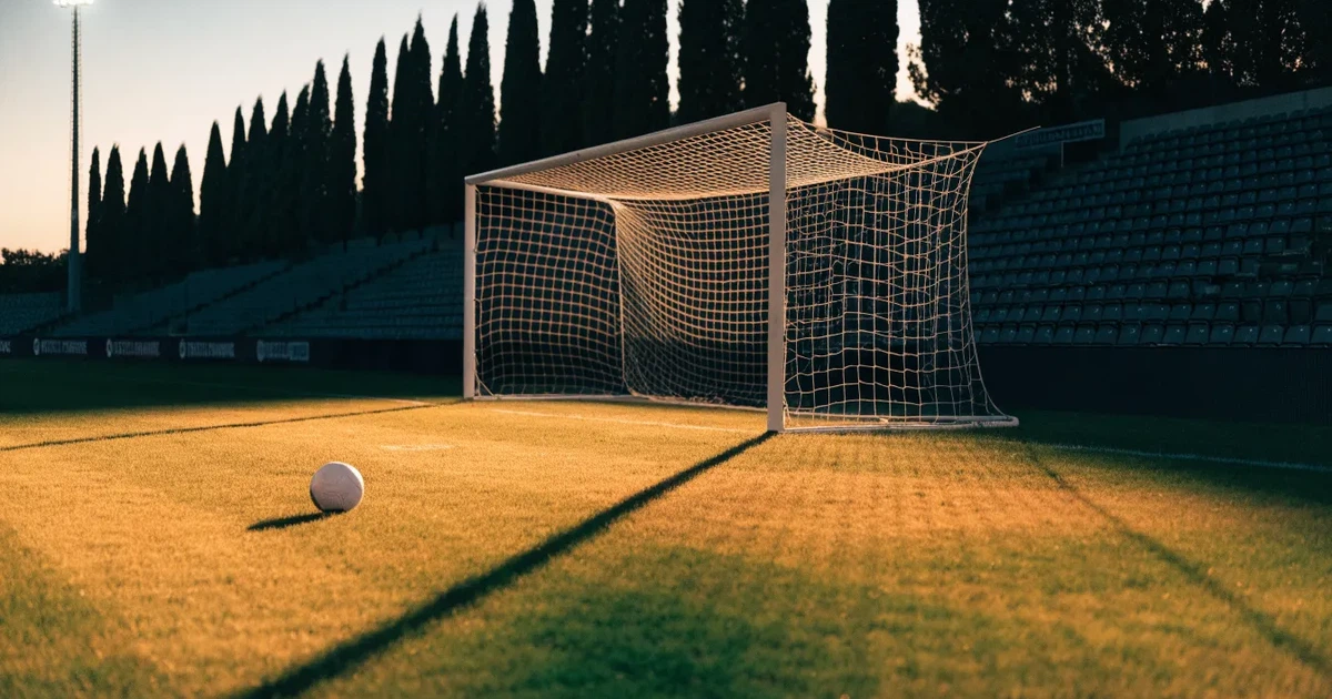 Ein Fußballtor mit Netz auf einem leeren Platz im Abendlicht, der Rasen frisch gemäht, im Hintergrund Zypressen.