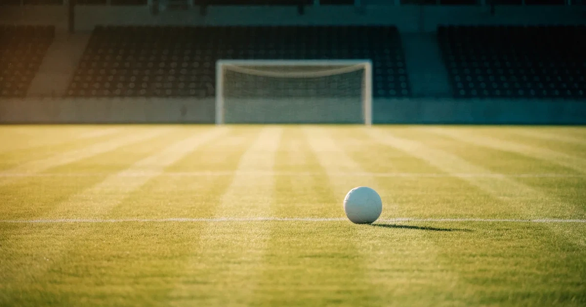 Ein Fußballplatz mit frisch gemähtem Rasen im Abendlicht, ein Ball liegt im Strafraum, im Hintergrund unscharf eine leere Tribüne.