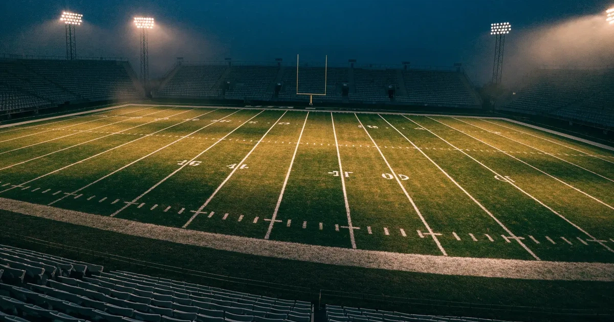 Ein American-Football-Feld mit weißen Yardlinien im Flutlicht, von einer hohen Tribüne aus gesehen, die Ränge leer, Zahlen nicht erkennbar.