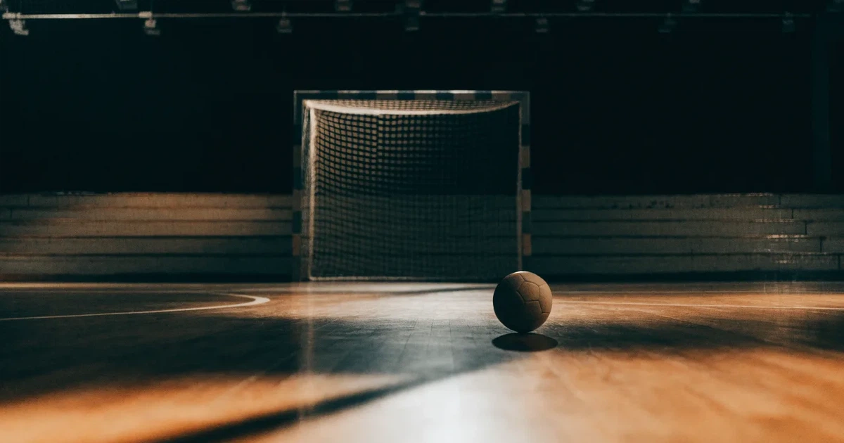 Ein Handball liegt auf dem Hallenboden vor einem Tor mit Netz, die leere Halle wird von Flutlicht beleuchtet.