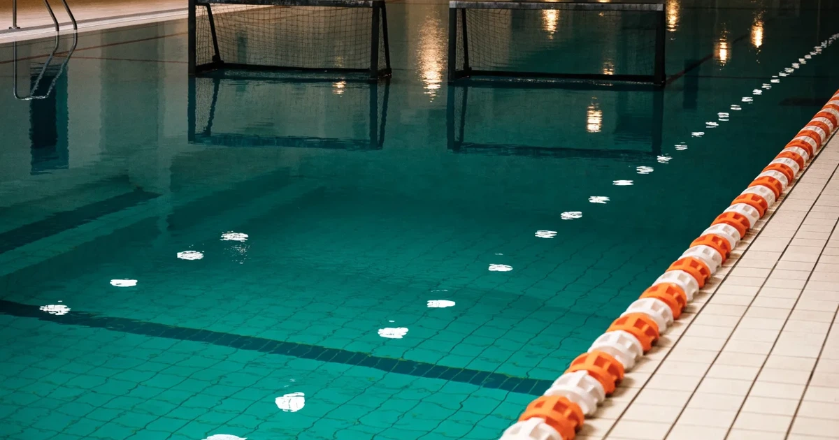Ein Schwimmbecken mit türkisfarbenem Wasser und zwei schwimmenden Torrahmen am gegenüberliegenden Beckenrand, menschenleer unter Hallenlicht.