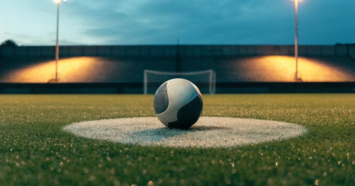 Ein Fußball liegt auf dem Anstoßpunkt eines leeren Rasenplatzes, im Hintergrund eine Tribüne und Flutlichtmasten in der Abenddämmerung.