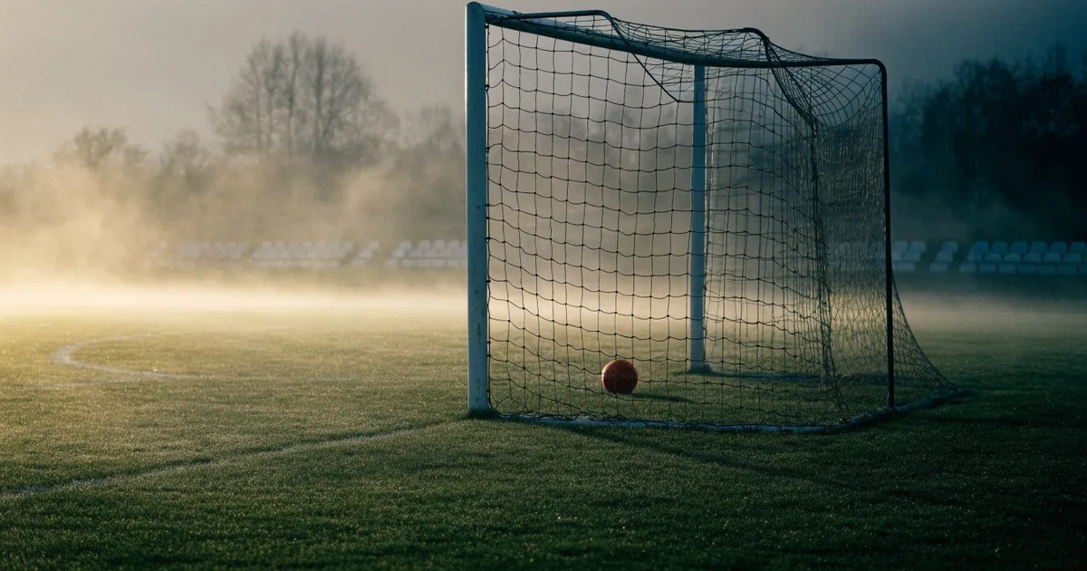 Ein Fußballtor mit Netz steht im Morgennebel auf einem leeren Rasenplatz, im Hintergrund die Silhouette einer kleinen Tribüne.