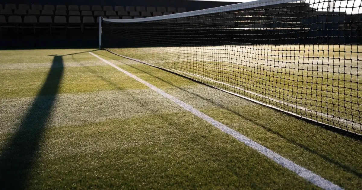 Ein Tennisplatz mit Rasenbelag im Morgenlicht, Netz und weiße Linien, im Hintergrund eine ruhige Tribüne.