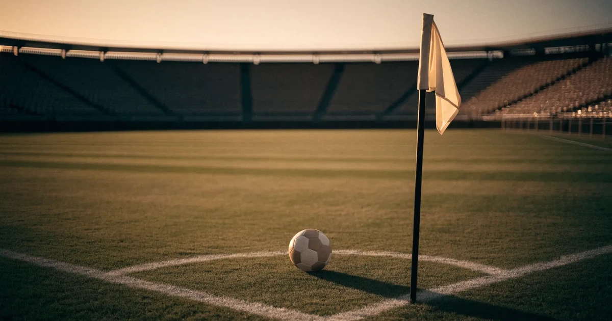 Ein Fußball liegt am Viertelkreis an der Eckfahne eines Rasenplatzes, im Hintergrund leere Tribünen in warmem Abendlicht.