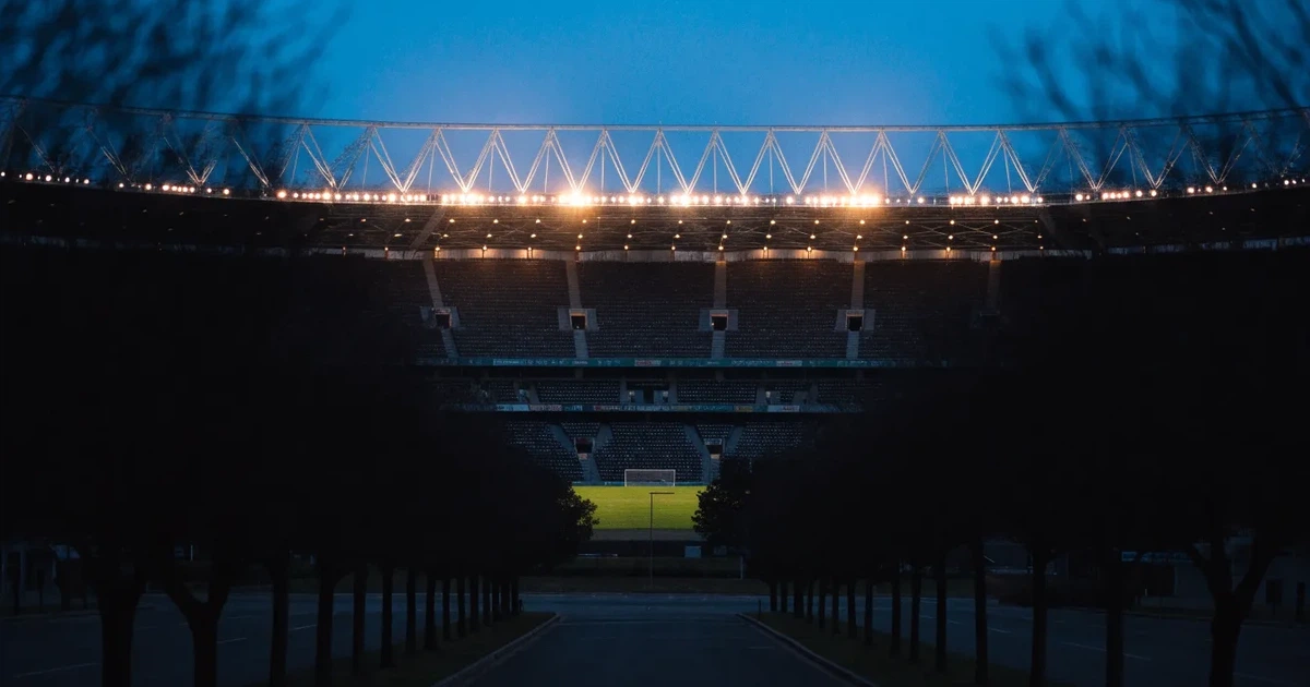 Ein Fußballstadion bei Nacht aus großer Entfernung, hell erleuchtet unter Flutlicht, davor eine dunkle Straße und Bäume.